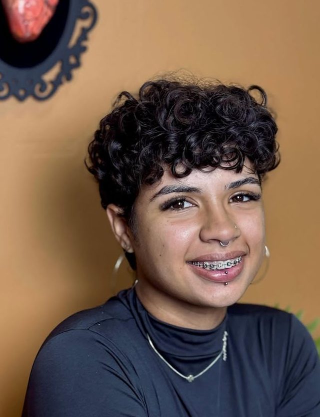 Cliente sorrindo, com piercing no septo e no lábio, em frente à parede laranja do studio.