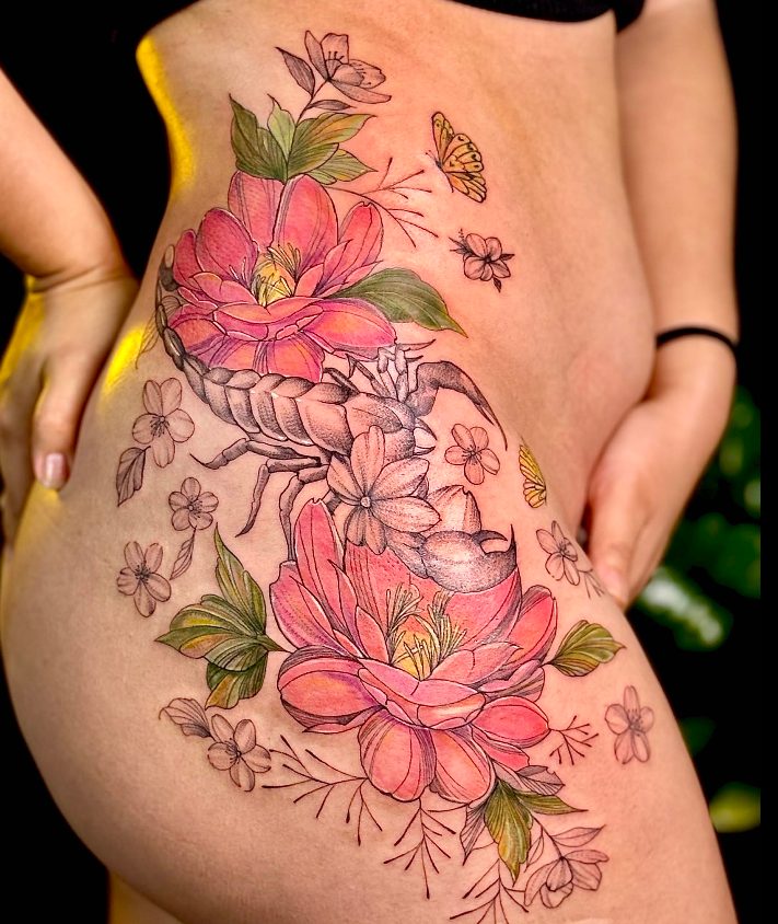 Tattoo colorida no quadril: flores cor-de-rosa com folhas verdes e um escorpião em preto.