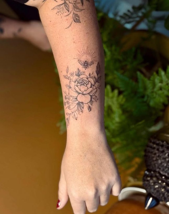 Tattoo em traço fino no antebraço: uma flor com folhas e uma abelha acima.