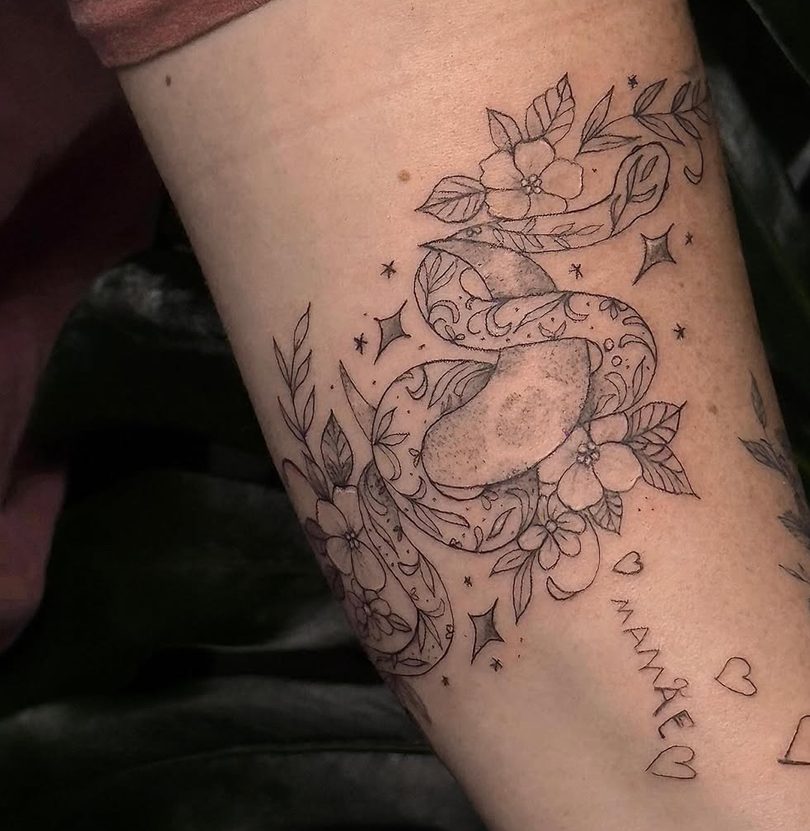 Tattoo em traço fino no braço: uma serpente entre flores, folhas e pequenas estrelas.