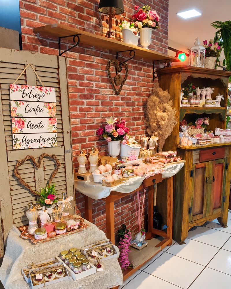 Interior do Armazém da Ju: parede de tijolos, veneziana antiga com uma placa, armário rústico e mesas com produtos e flores.
