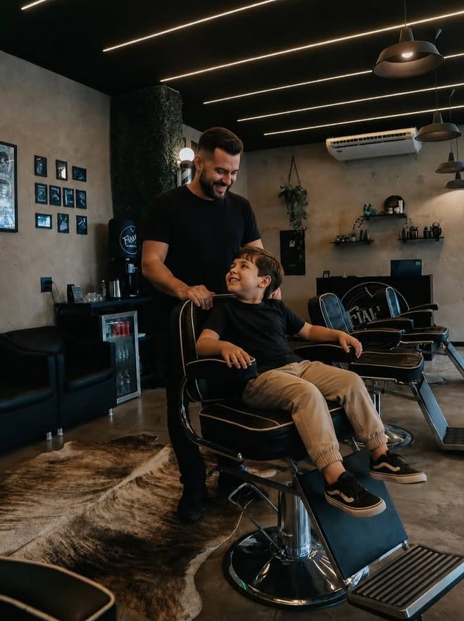 Barbeiro e um menino sentado na poltrona da barbearia, os dois sorrindo um para o outro.
