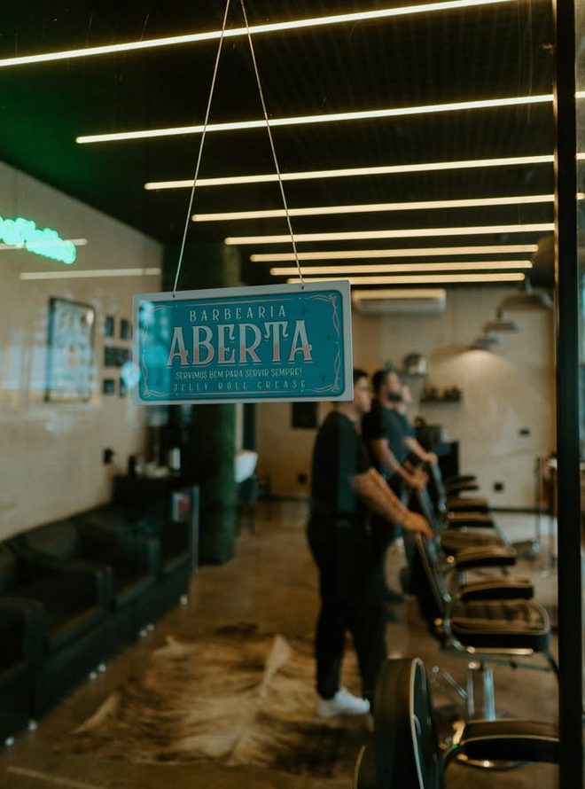 Placa pendurada na porta com os dizeres Barbearia Aberta, servimos bem para servir sempre. Ao fundo, barbeiros atendendo.