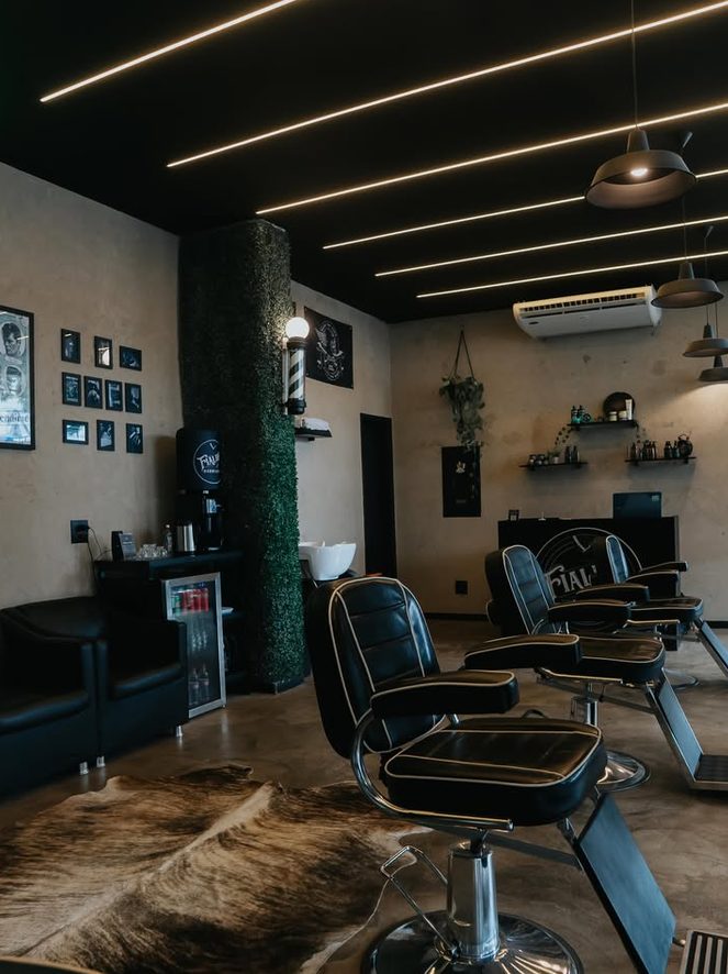 Salão da Fialho Barbearia: poltronas de barbeiro pretas, tapete de couro no piso, poste de barbeiro na parede e iluminação em linhas no teto.