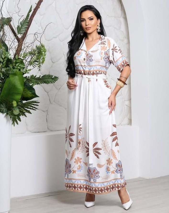 Mulher com vestido longo branco de estampa floral em tons terrosos e azul, com cinto.