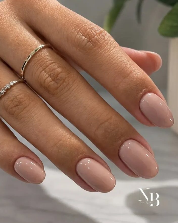 Mãos com unhas em gel nude, curtas e brilhantes, e anéis finos dourados.