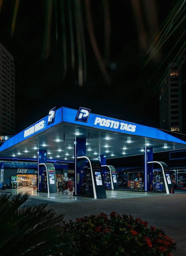 Posto Tacs à noite: cobertura azul iluminada com o nome do posto, bombas de combustível e a conveniência ao fundo.