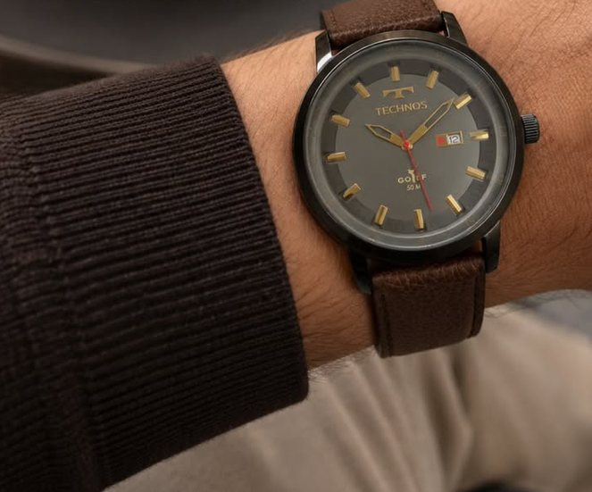 Relógio com pulseira de couro marrom e mostrador escuro, no pulso, sob um casaco de lã.