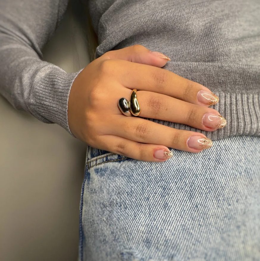 Mão apoiada no jeans, com unhas alongadas em tom natural e pontas com brilho dourado.