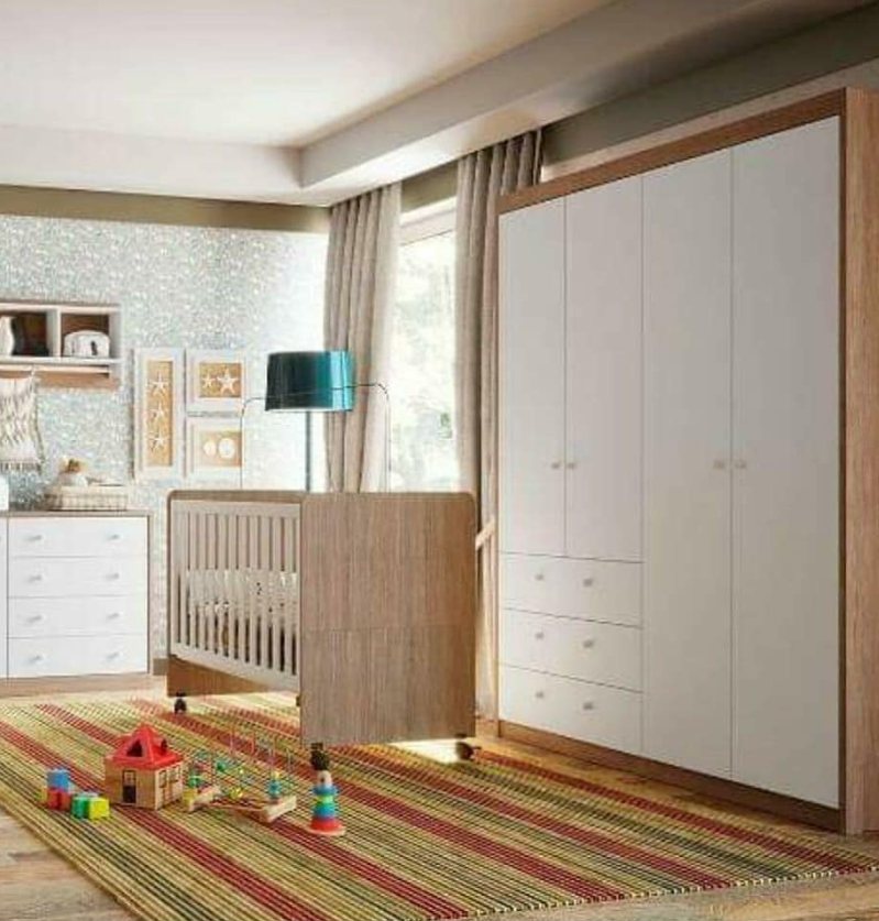 Quarto com guarda-roupa branco de quatro portas, berço em madeira e cômoda, com brinquedos sobre um tapete listrado.